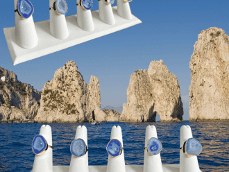 Display of dark blue geode rings on white holders, with a scenic background of the Faraglioni rock formations emerging from the blue sea under a clear sky.