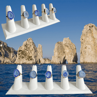 Display of dark blue geode rings on white holders, with a scenic background of the Faraglioni rock formations emerging from the blue sea under a clear sky.