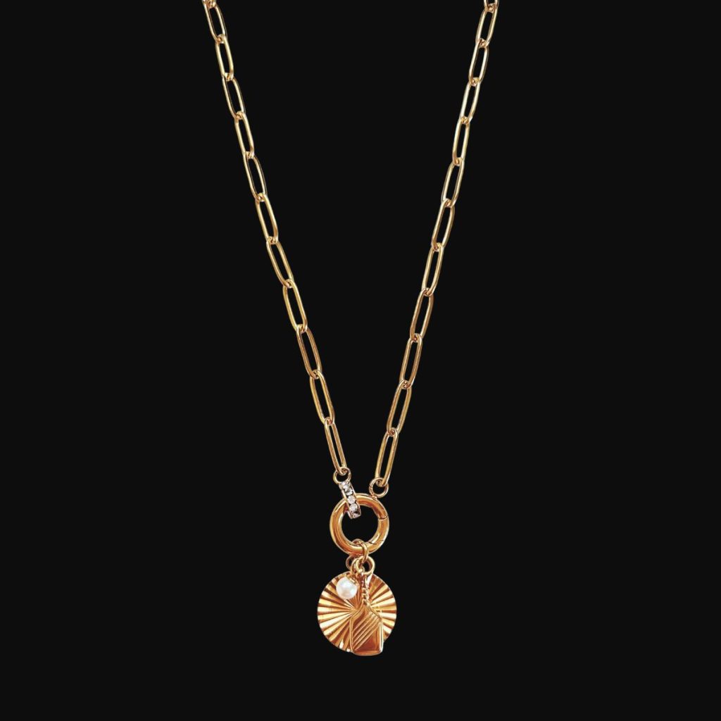 Gold paperclip link chain necklace featuring a circular spring clasp holding a round fluted sunburst disc charm, a freshwater pearl drop, and a small textured paddle tag on a black background.