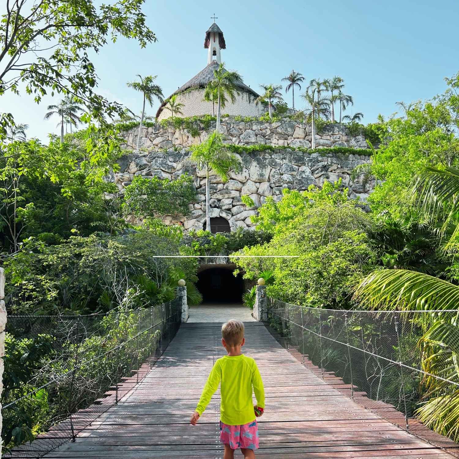A small boy walking across a wooden suspension bridge surrounded by lush green jungle leaves toward the hillside chapel at Hotel Xcaret Mexico.