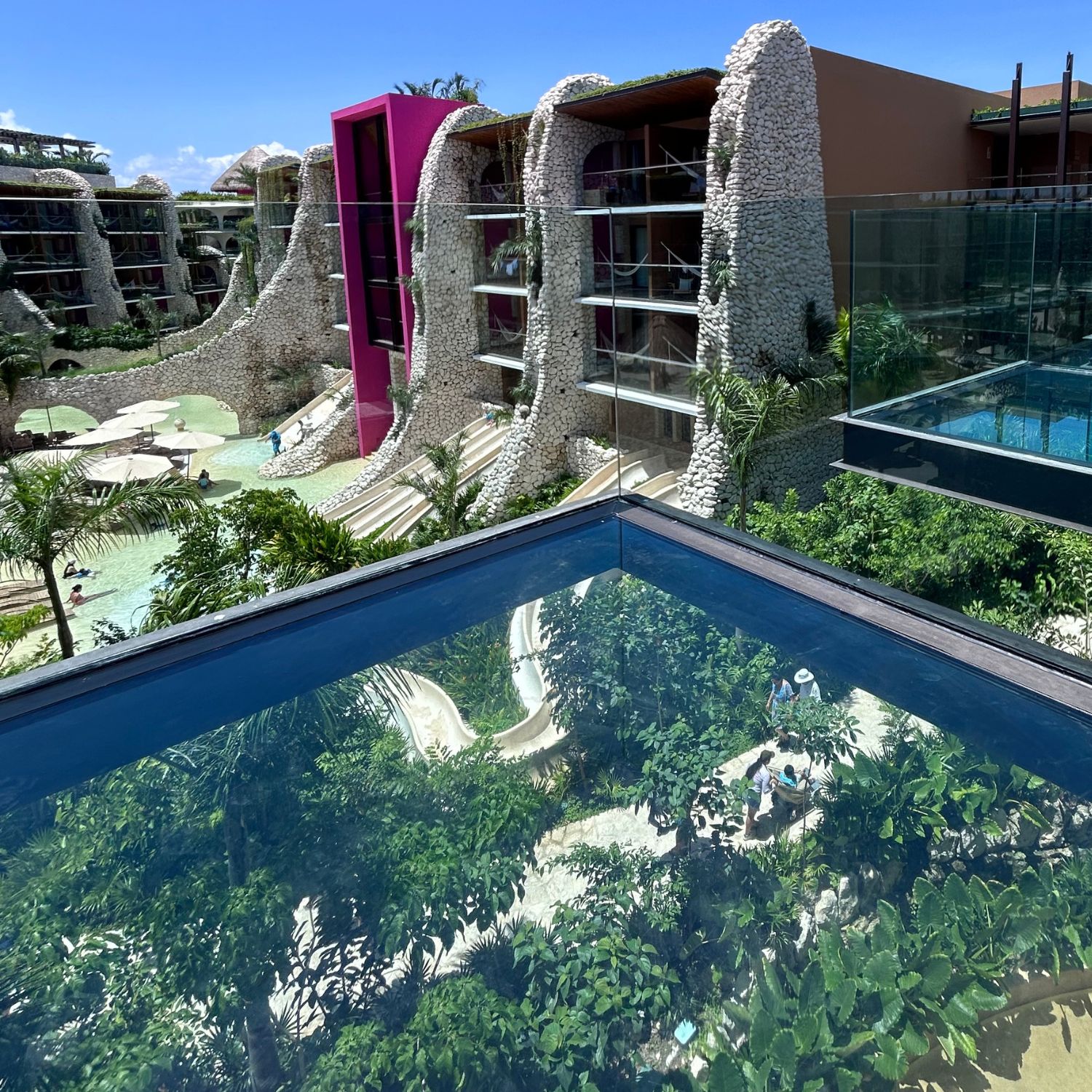 An exterior view of the modern jungle suites at Hotel Xcaret Mexico, showcasing the resort Casas, family waterslides, and a transparent glass balcony over the water.