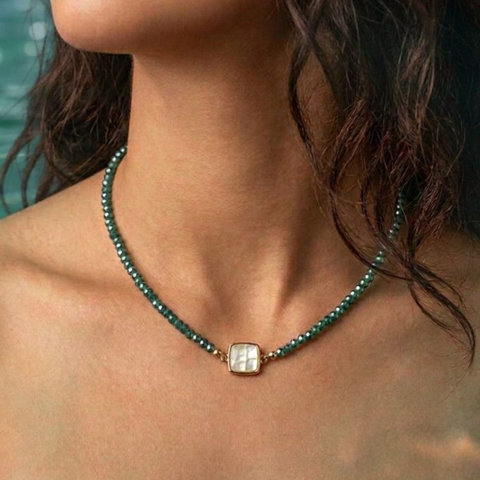 Close-up of a square mother of pearl pendant necklace on a delicate green beaded chain worn by a woman with dark wavy hair and tan skin.