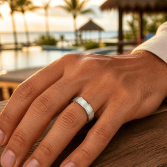 A close-up of a man's hand wearing a silver stainless steel wedding band with a brushed center and polished edges, resting on a wooden surface with a tropical resort and beach sunset blurred in the background.