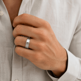 A close-up lifestyle shot of a man's hand adjusting his beige linen button-down shirt, showing a silver dual-finish stainless steel band with a recessed brushed center and polished outer edges on his ring finger.