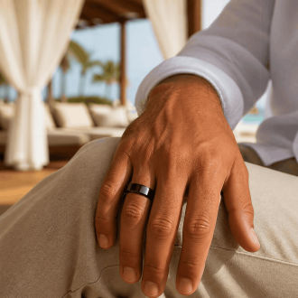 A close-up lifestyle shot of a man's hand resting on his knee while wearing a glossy black beveled wedding band, paired with khaki trousers and a white linen shirt at a luxury resort.