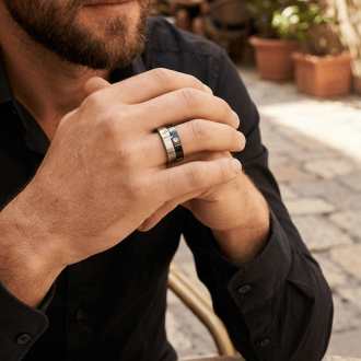 A close-up of a man's clasped hands showing a two-tone industrial ring with a brushed silver band and a polished black inlay featuring screw details, worn with a black button-down shirt at an outdoor café.